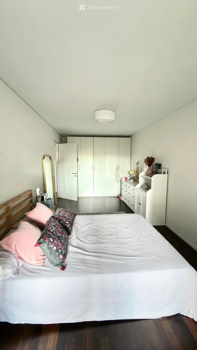 A modern bedroom with a large bed, striped bedding, pink and patterned pillows, a wooden headboard, white wardrobe, a gold-framed mirror, and a nightstand with a lamp and decorative items.