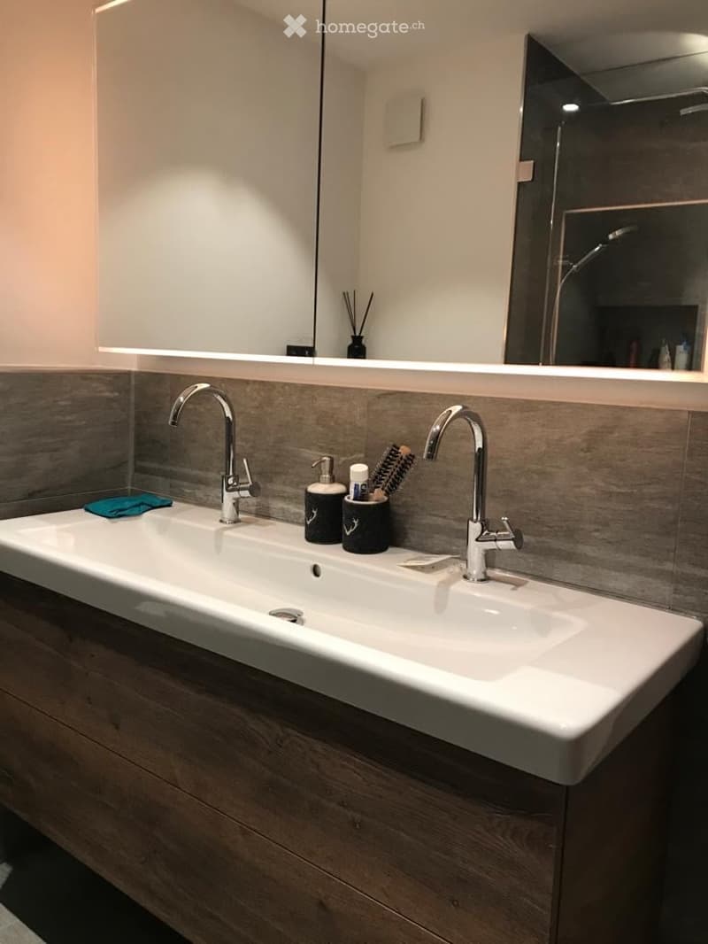 Modern bathroom with a double sink, sleek chrome faucets, a large mirror, and a wooden vanity. Personal care items are neatly arranged on the sink counter.