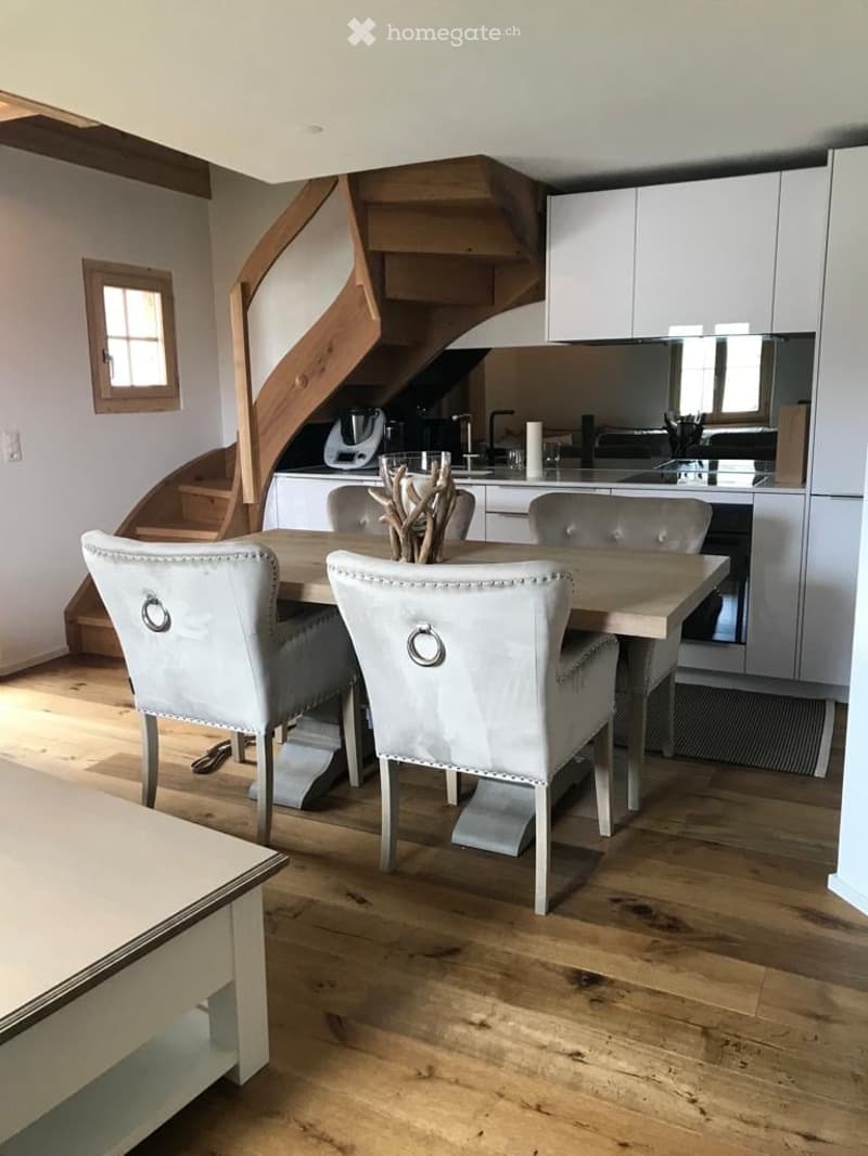 Dining area with a wooden table and four upholstered chairs, adjacent to a modern kitchen with white cabinets, stainless steel appliances, and a staircase leading to an upper level.