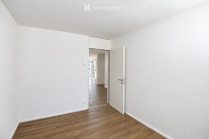Empty room with white walls and wooden flooring, featuring an open doorway leading to another room.