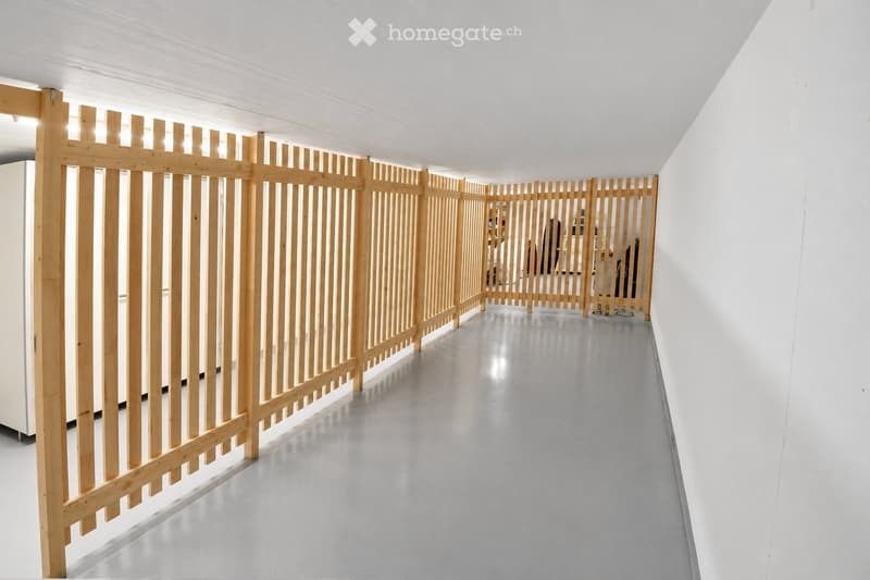 A modern, minimalist hallway with white walls and a smooth grey floor. Wooden slatted partitions line the corridor, creating a sense of separation and privacy.