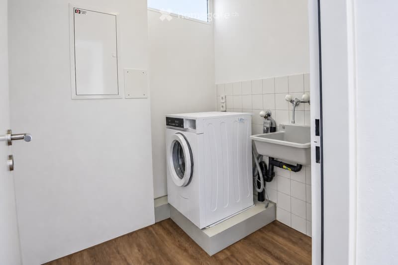 Modern bathroom with a white washing machine and a sink, white tiled walls, and wooden flooring.