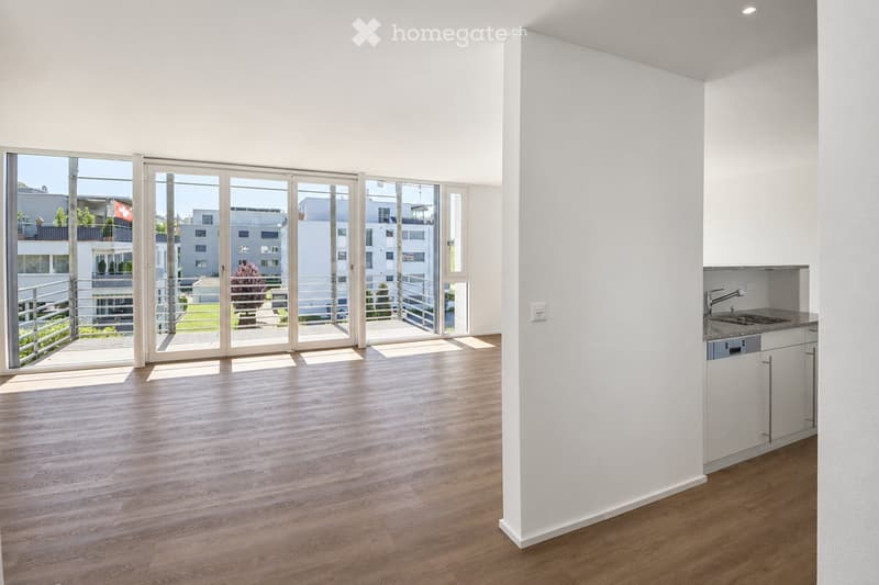 Bright, unfurnished room with wooden flooring, large sliding glass doors leading to a balcony, and a small kitchenette area with a sink and cabinets.