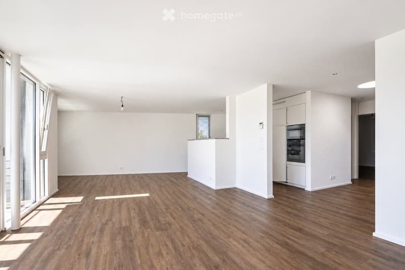 Bright, empty room with wooden flooring, white walls, and large windows allowing natural light. The space includes a small kitchen area with white cabinets and a built-in oven.