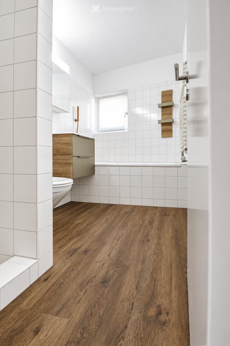 Modern bathroom with white tiled walls, wooden flooring, a toilet, a sink with a mirror, a bathtub, and a shower rack.