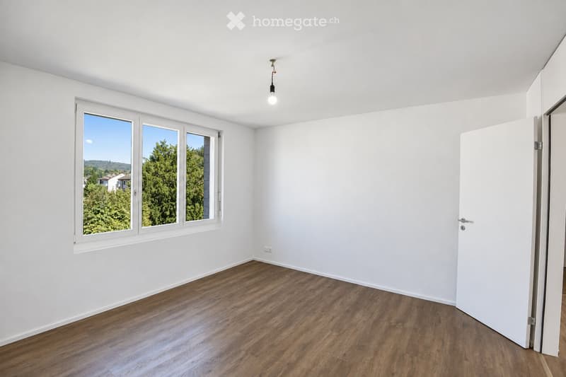 Empty room with white walls, wooden flooring, and large windows showing greenery outside. No furniture or equipment present.
