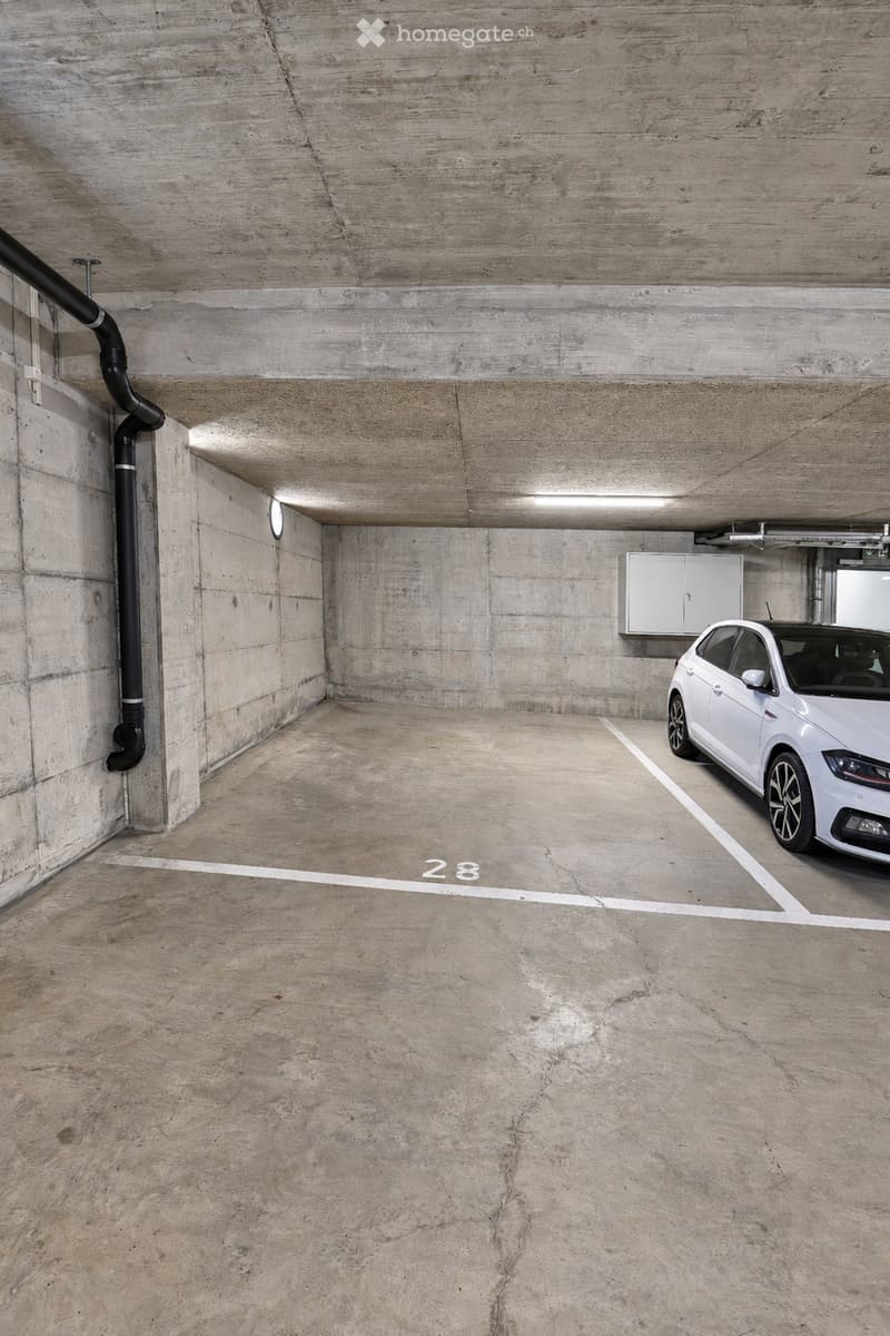 Underground parking space with a single white car parked in a marked bay, concrete walls and ceiling, and a numbered parking spot.