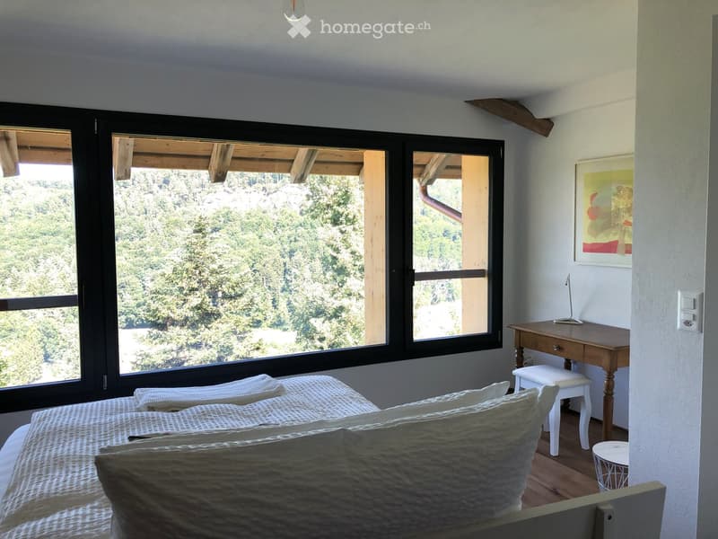 Bright bedroom with large windows offering a scenic view, featuring a bed with pillows, a wooden desk with a stool, and a lamp on the desk.