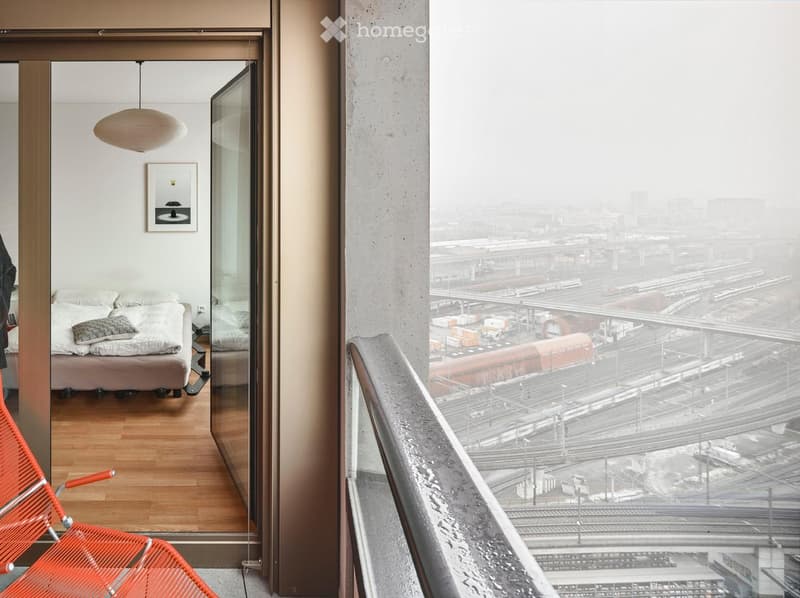 Modern bedroom with a bed, a red plastic chair, a pendant light, and a picture frame on the wall, overlooking a train station.