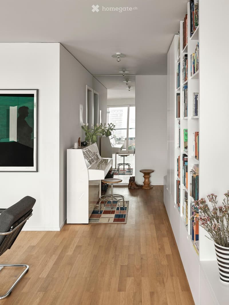 Modern hallway with wooden flooring, white walls, built-in bookshelf, and a small seating area with a piano and chairs.