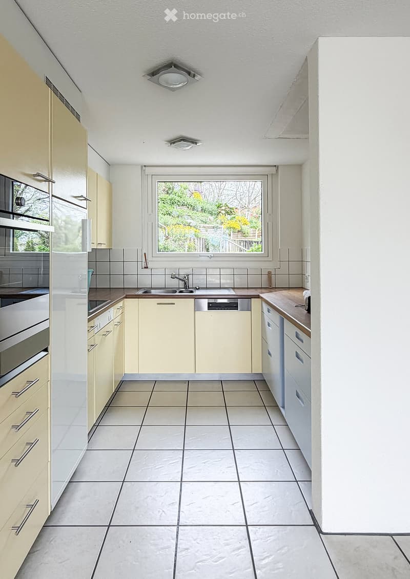 Bright kitchen with light-colored cabinets, a central island, and a large window overlooking a garden. Features include a sink, dishwasher, and oven.
