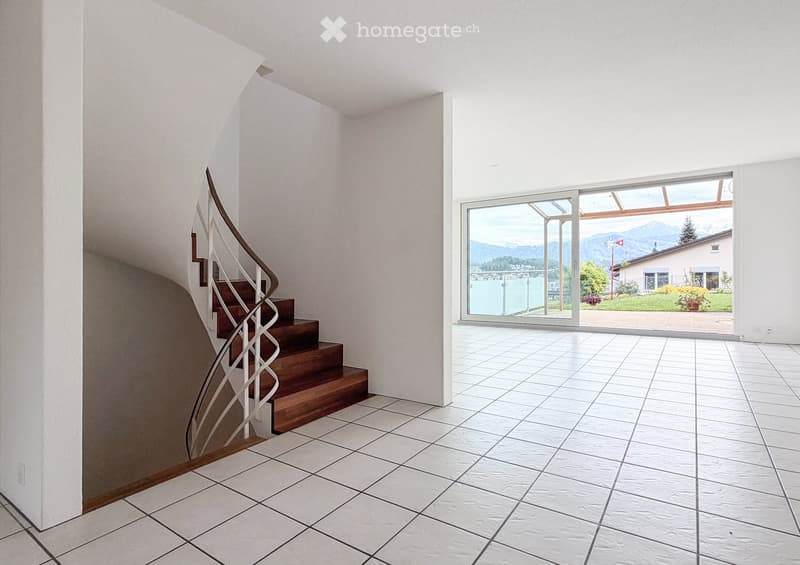 Bright hallway with white tiled flooring, a wooden staircase with a modern metal railing, and a large glass door leading to a scenic outdoor view.