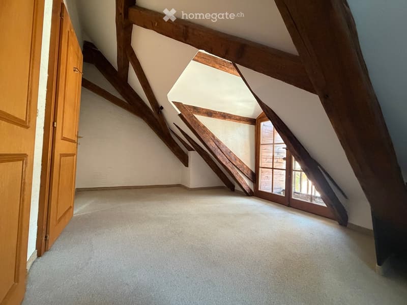An empty attic space with exposed wooden beams, carpeted floor, and a large window with wooden frame. The room has a wooden door on the left side.