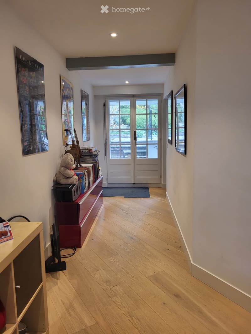 A well-lit hallway with wooden flooring, white walls, and several framed pictures. A small table with a teddy bear and books is placed on the left side, and a large double door leads to an outdoor area.