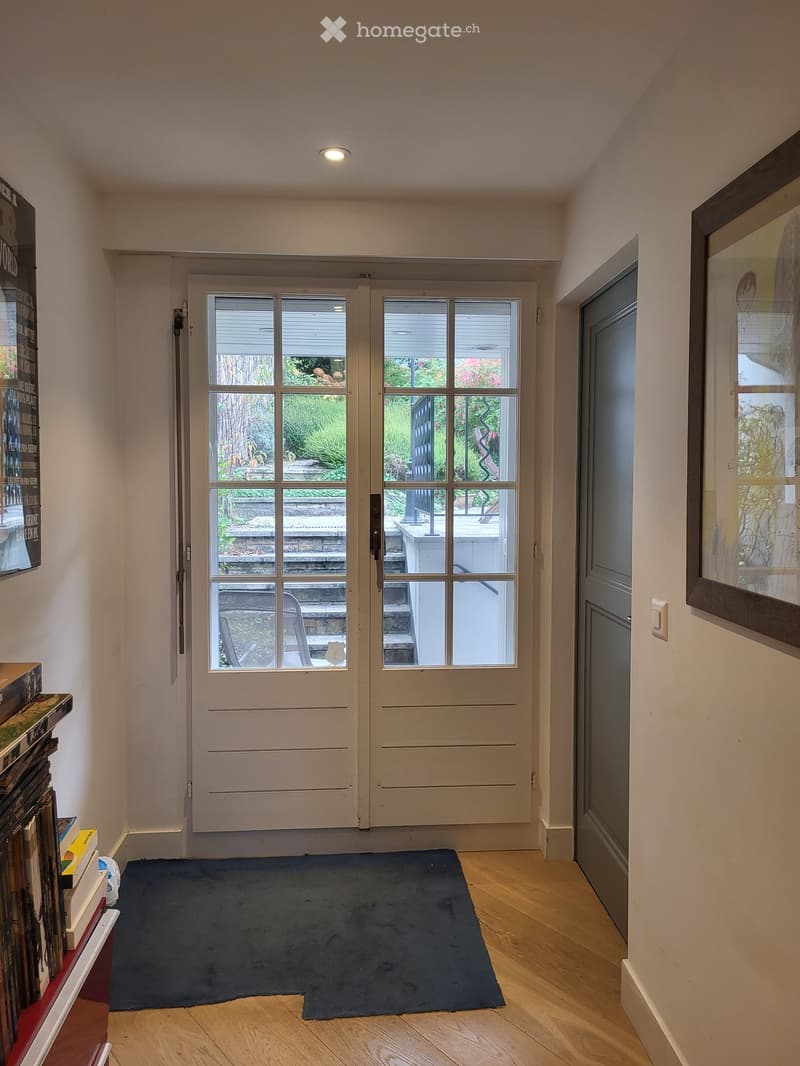 Hallway with wooden flooring, a dark blue rug, double glass doors leading to an outdoor area, and a stack of books and magazines on a shelf to the left.