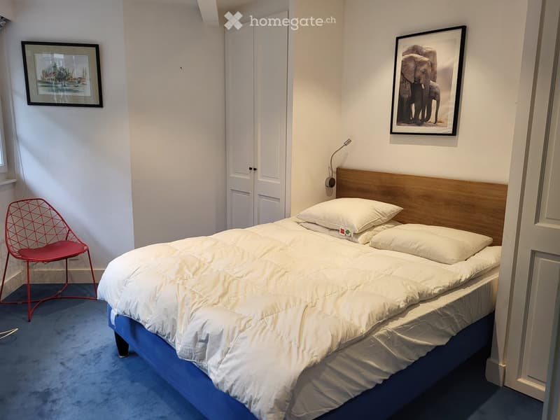 Bedroom with a neatly made double bed, a red wire chair, and two framed pictures on the wall.