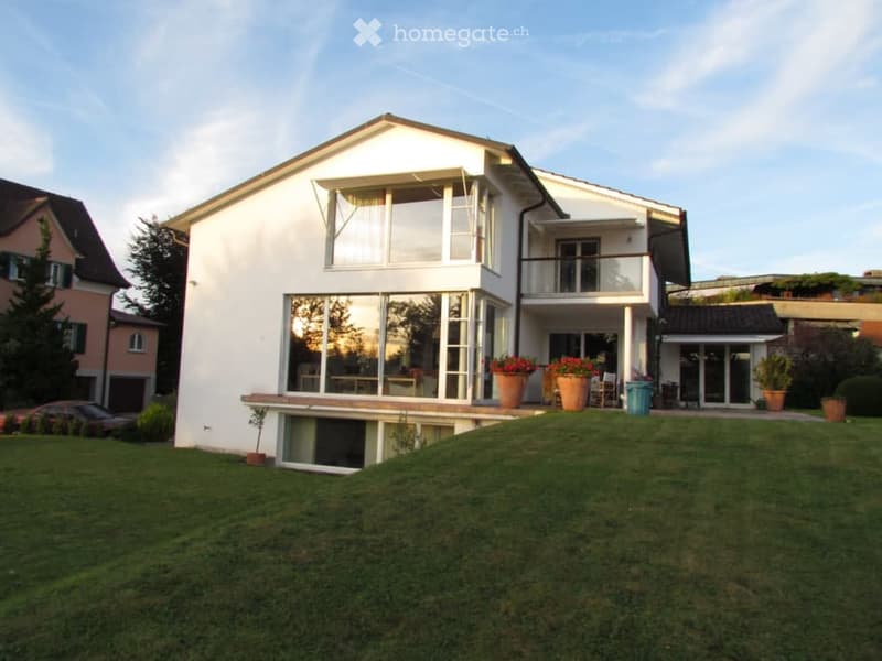 modern 2-story house, white exterior, large windows, balcony, raised ground floor, surrounded by a large lawn and greenery