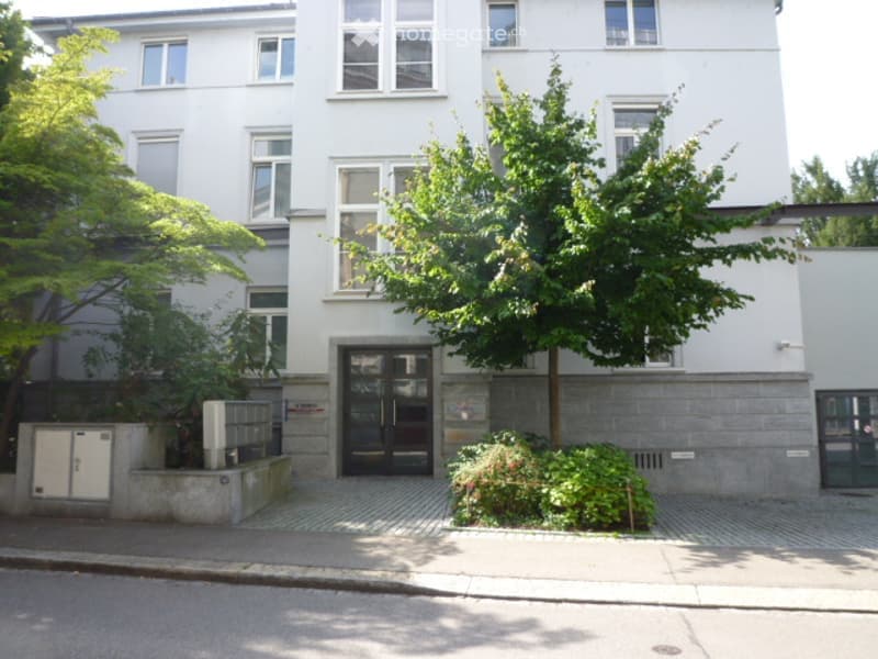 Modern multi-story building with white facade, large windows, and a small landscaped area in front.