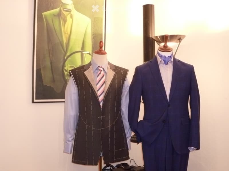 Two mannequins dressed in formal suits, one in a vest and tie, the other in a full suit, with a lamp and framed picture in the background.