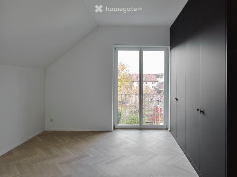 Empty room with wooden floor, white walls, large sliding glass door leading to balcony, and built-in dark grey wardrobe.