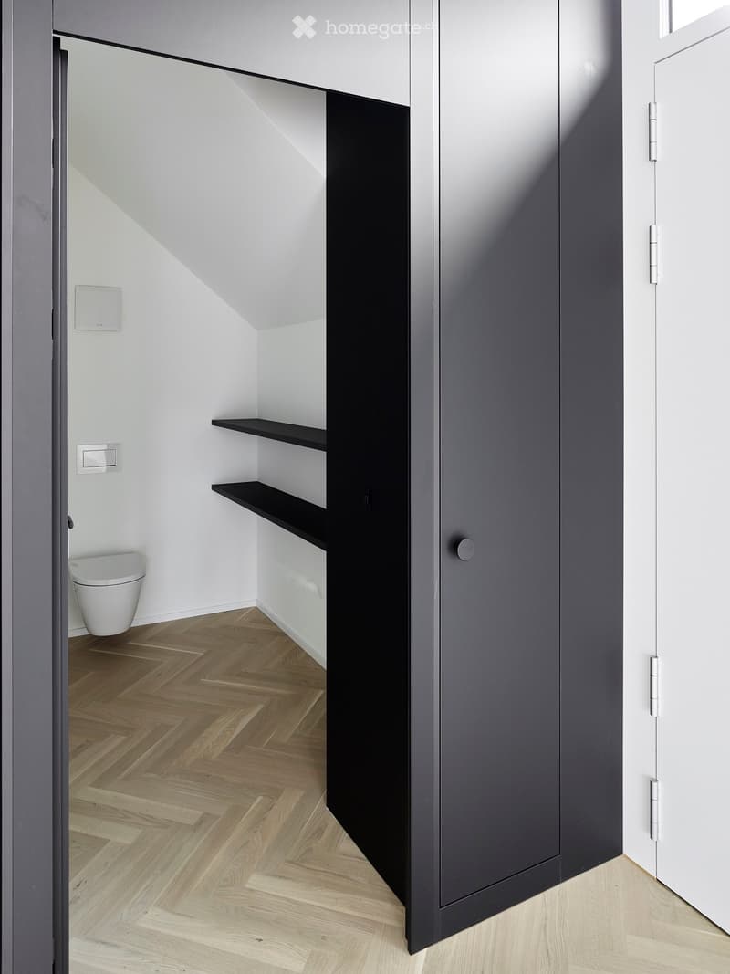 Modern bathroom with white walls, wooden herringbone floor, toilet, and black shelving unit.