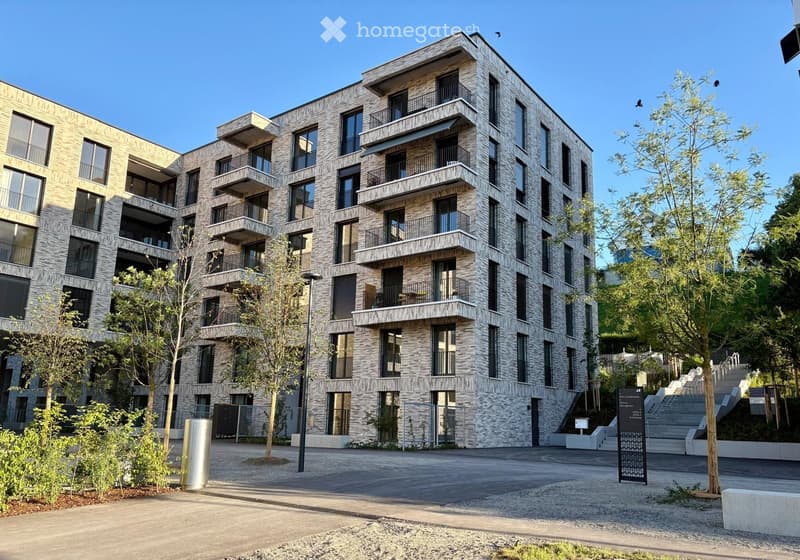 Modern multi-story residential building with balconies, large windows, and a stone facade, surrounded by trees and a paved area.