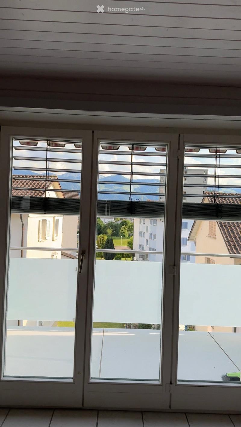 Empty room with large windows and blinds, overlooking a view of mountains and buildings. The floor is tiled, and the walls are painted white.