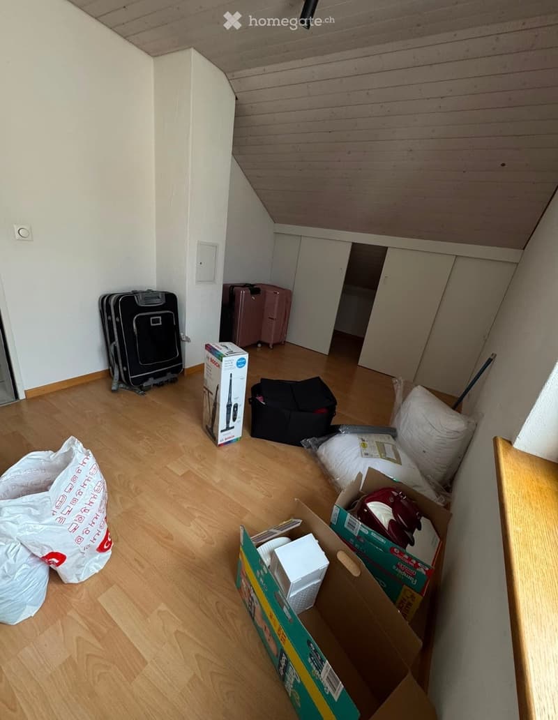 Empty attic room with wooden flooring, white walls, and exposed wooden ceiling beams. Various boxes, suitcases, and bags are scattered around the space.