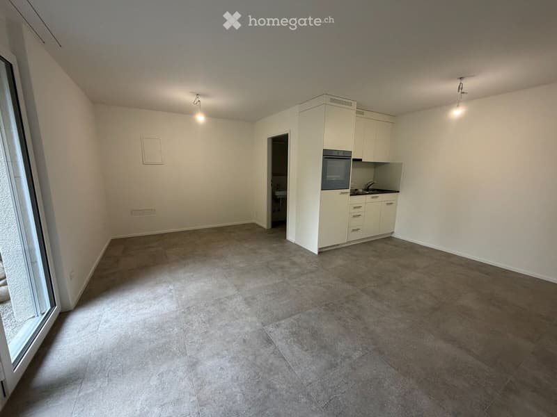 Empty room with tiled floor, white walls, and a small kitchen area with cabinets and appliances visible in the background.
