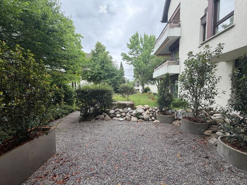 Garden area with gravel pathway, potted plants, rocks, and a modern building with a balcony in the background.