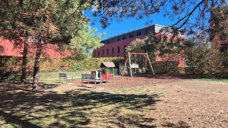 Outdoor playground area with swings, a small playhouse, benches, and trees, surrounded by a red brick building.