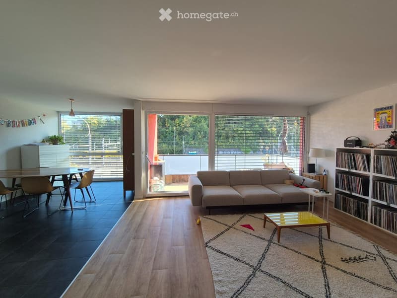 Modern living room with a sofa, coffee table, dining table, chairs, bookshelf, and a lamp. Large windows and a balcony are visible.