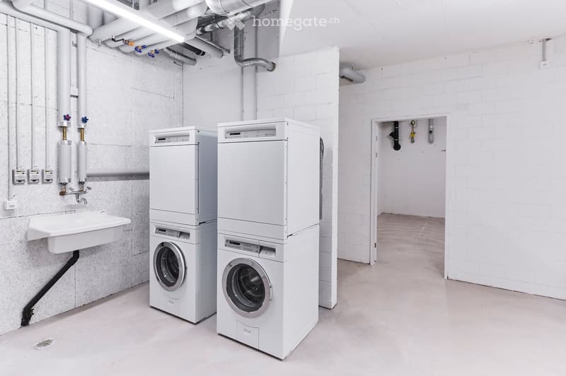 A clean, modern laundry room with white brick walls, a concrete floor, and two stacked washing machines and dryers. A small sink is visible on the left side.