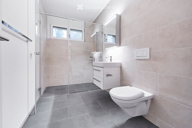 Modern bathroom with beige tiled walls, grey tiled floor, white sink, toilet, and a mirrored cabinet with built-in lighting above the sink. A glass-enclosed shower area is visible in the background.