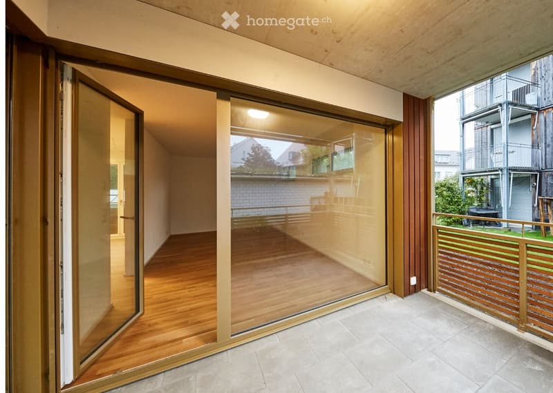 Modern balcony with large glass sliding doors, wooden flooring inside, tiled flooring outside, wooden railing, and view of neighboring buildings and greenery.