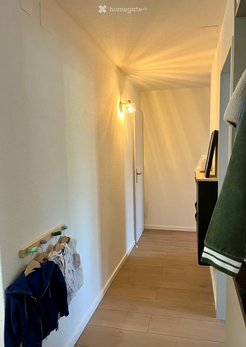 A narrow hallway with light wooden flooring, white walls, and a coat rack holding jackets. A small table with a picture frame is visible at the end of the hallway.