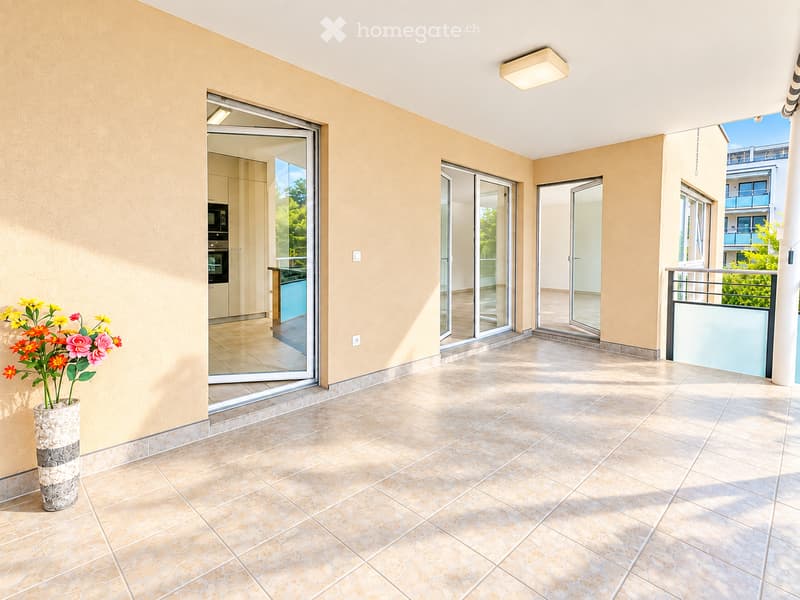 Balcony with tiled floor, beige walls, sliding glass doors leading to interior, metal railing, and a decorative vase with flowers on the left side.