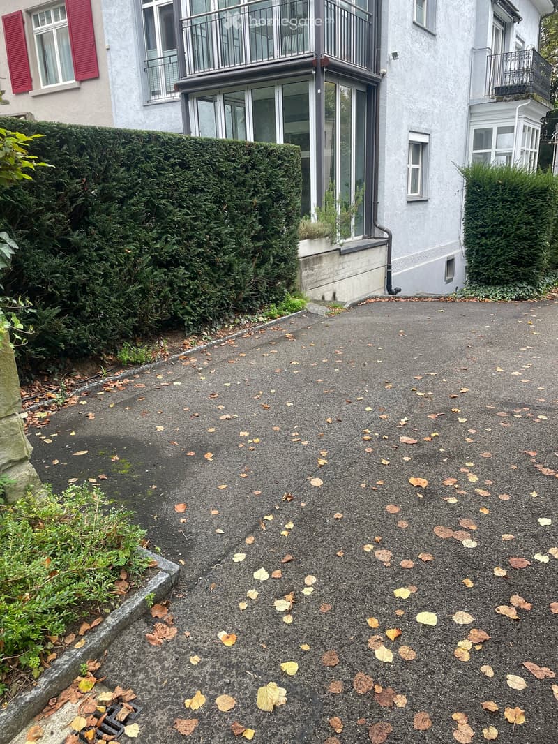 House with red shutters, balconies, glass doors, asphalt driveway, hedges, leaves scattered on the ground