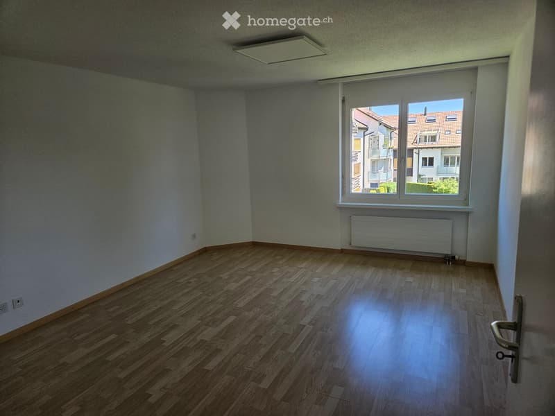 Empty room with wooden flooring, light-colored walls, and a large window offering a view of neighboring buildings.