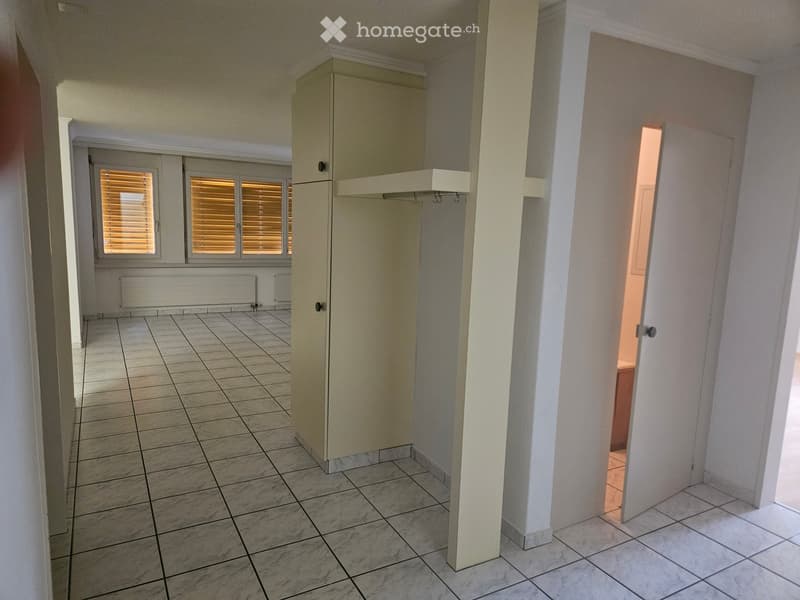 Empty hallway with tiled flooring, white walls, and a built-in closet with a shelf. A door leads to another room with warm lighting.