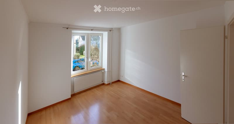 Empty room with wooden flooring, white walls, and a window with a view of a parked car and garden outside.