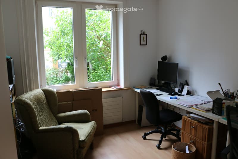 A furnished room with a sofa, desk, computer, and various office supplies. The room has wooden flooring and large windows with a view of greenery outside.