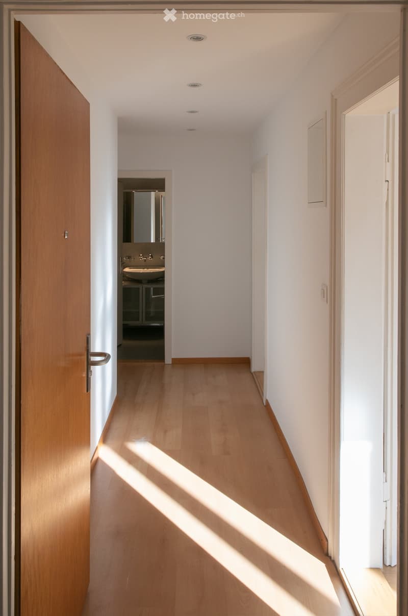 Bright hallway with wooden flooring, white walls, and recessed lighting. A wooden door is open on the left, revealing a bathroom with a sink and mirror. Sunlight streams in from the right side.