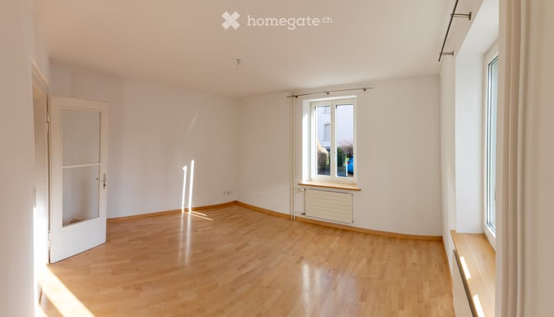 Empty room with wooden flooring, white walls, and large windows allowing natural light. Features a radiator and a door leading to another space.