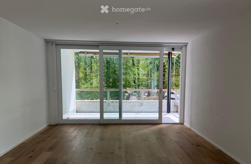 Empty room with wooden flooring and large sliding glass doors leading to an outdoor area with trees and parked cars visible.