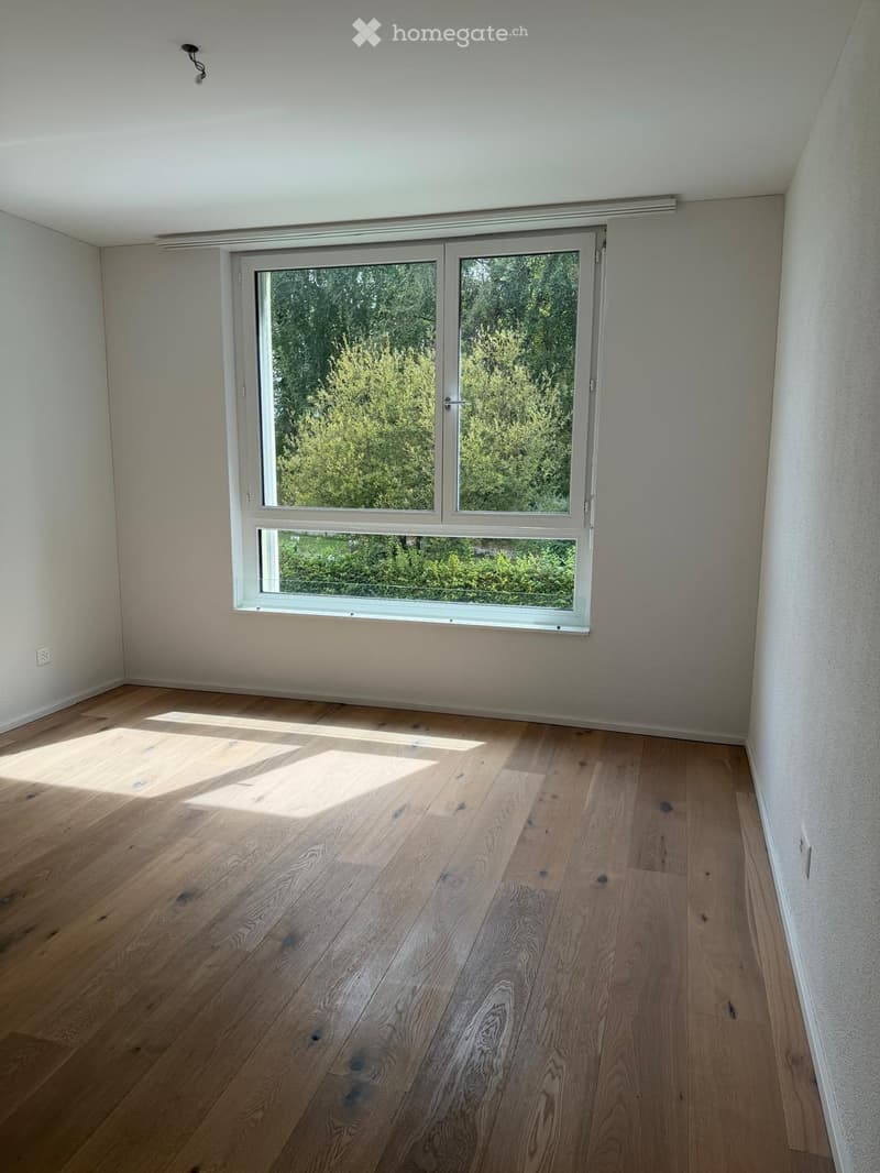 Empty room with light wooden flooring and large window showing greenery outside. Bare walls and no furniture.