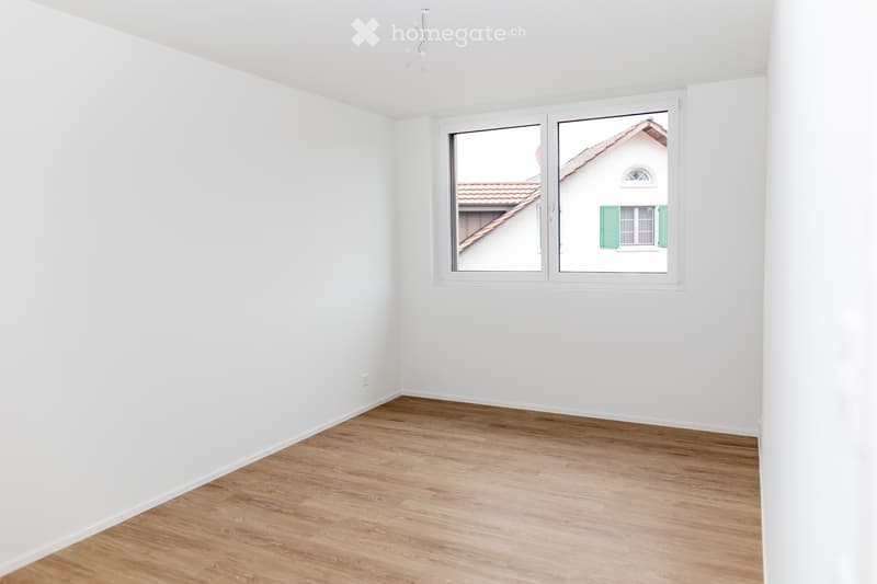Empty room with white walls and wooden flooring, featuring a large window with a view of a neighboring building.