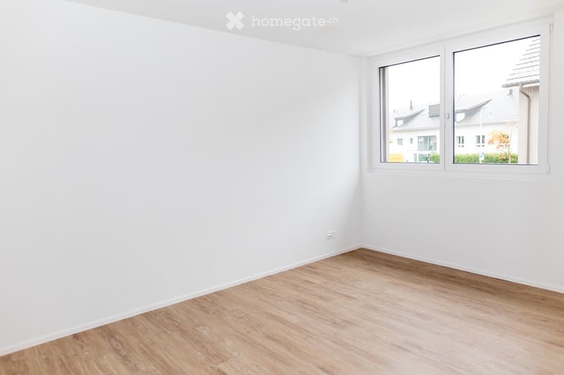 Empty room with white walls and light wooden flooring, featuring large windows with white frames overlooking a residential area.