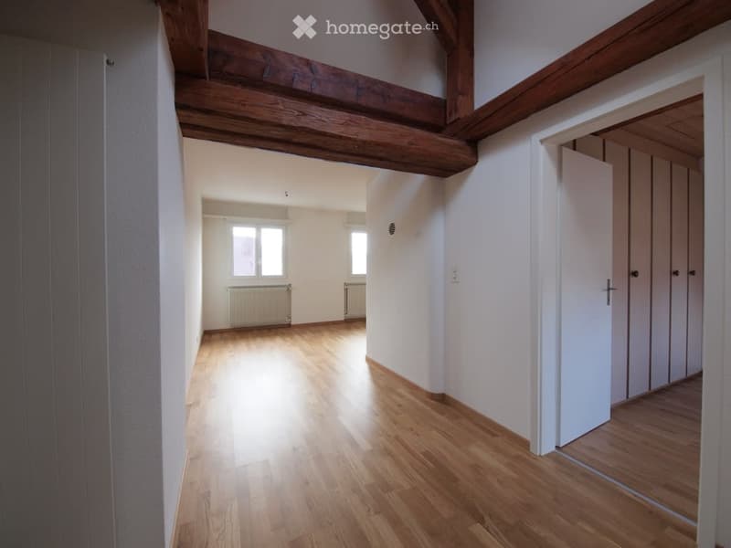 Empty room with wooden flooring, white walls, exposed wooden beams on the ceiling, and a window with a radiator below it.