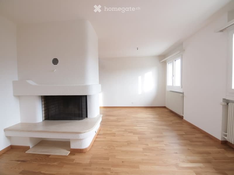 Empty room with light wooden flooring, white walls, and a built-in fireplace. Large windows allow natural light to fill the space.